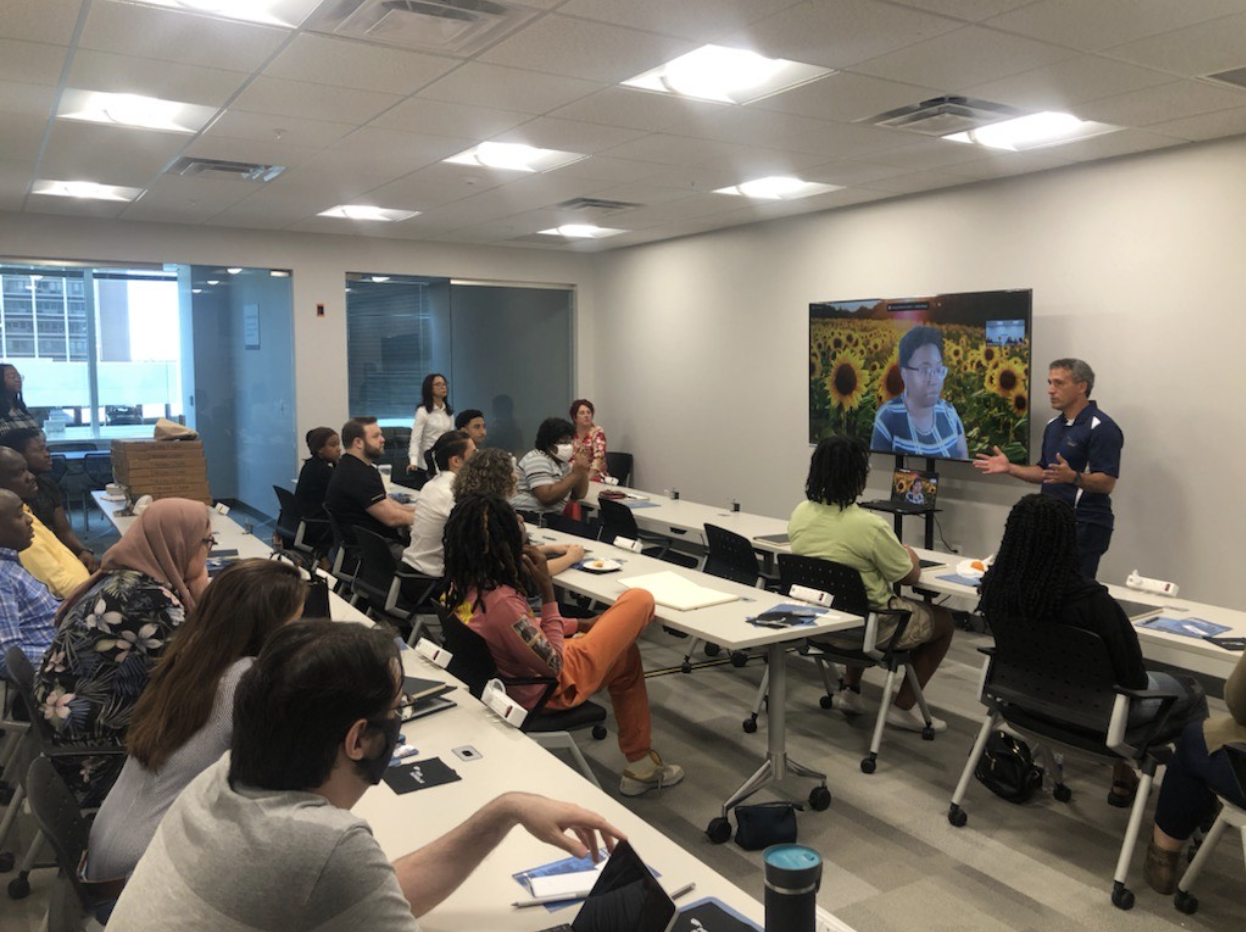 Adult learners seated in a classroom listening to an instructor at the front with a person appearing on a large screen.