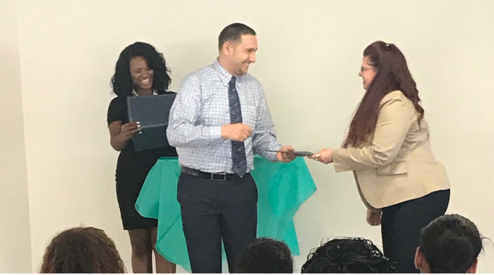 Three people on a stage exchanging certificates while an audience watches, capturing a graduation or award presentation.