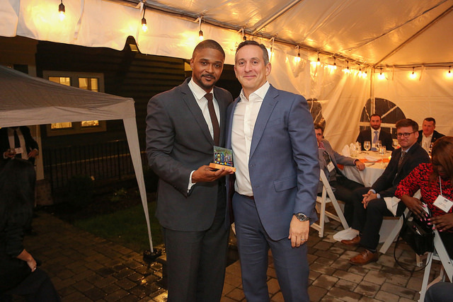 Two men in suits stand under a tent at a formal outdoor evening event, one holding a small award while guests sit at tables behind them.