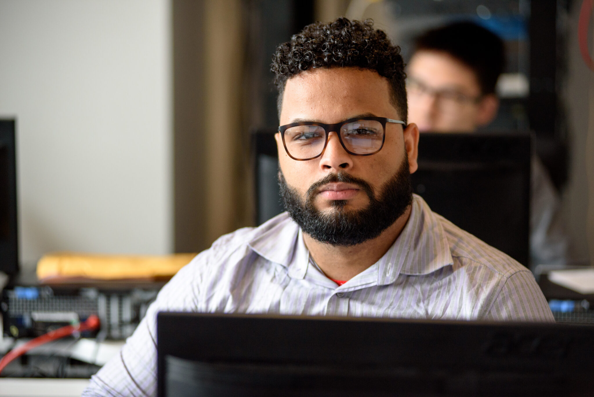 Young man with glasses and beard focused at a computer in a classroom.