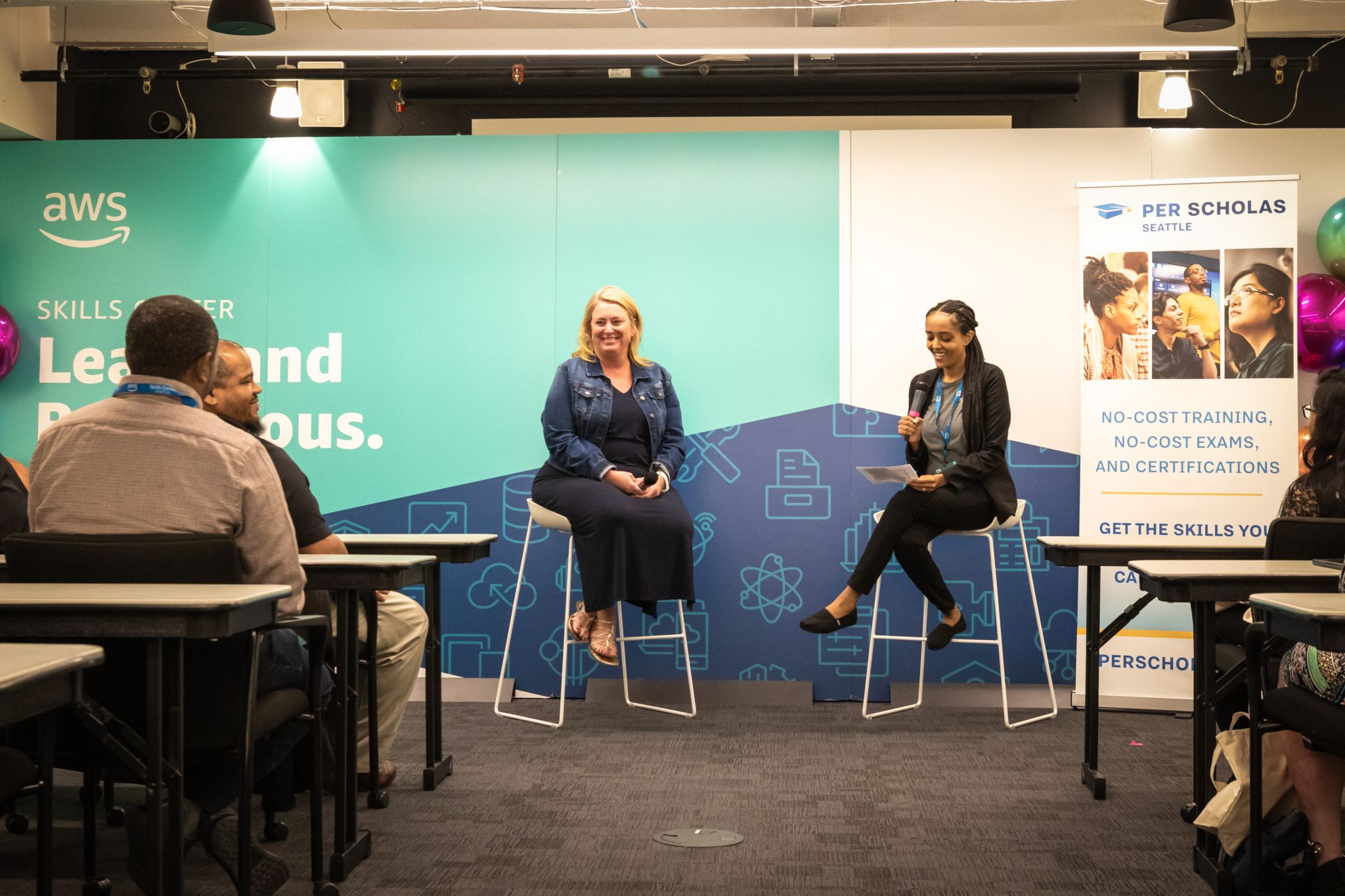 Two instructors on stools speaking to seated students at a Per Scholas and AWS skills event.