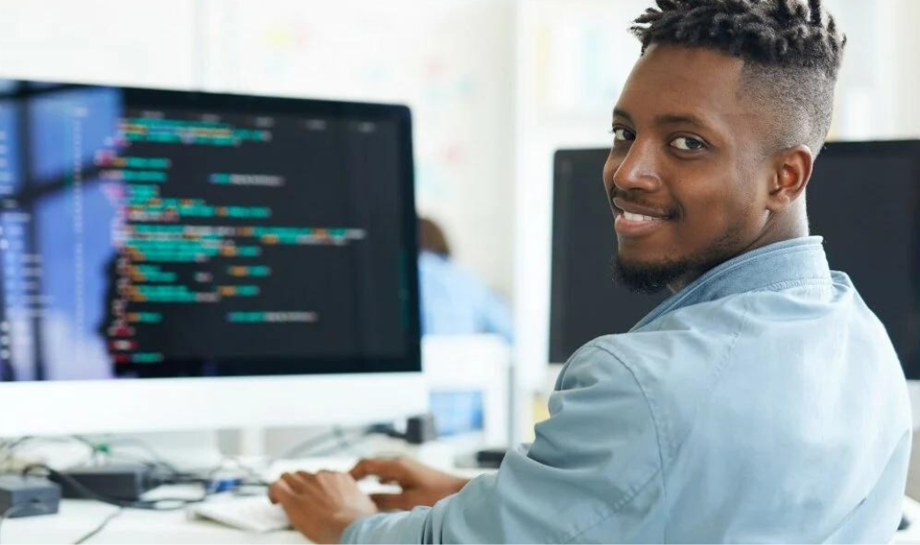Young man at a computer smiling over his shoulder while coding on a large monitor in an office setting.