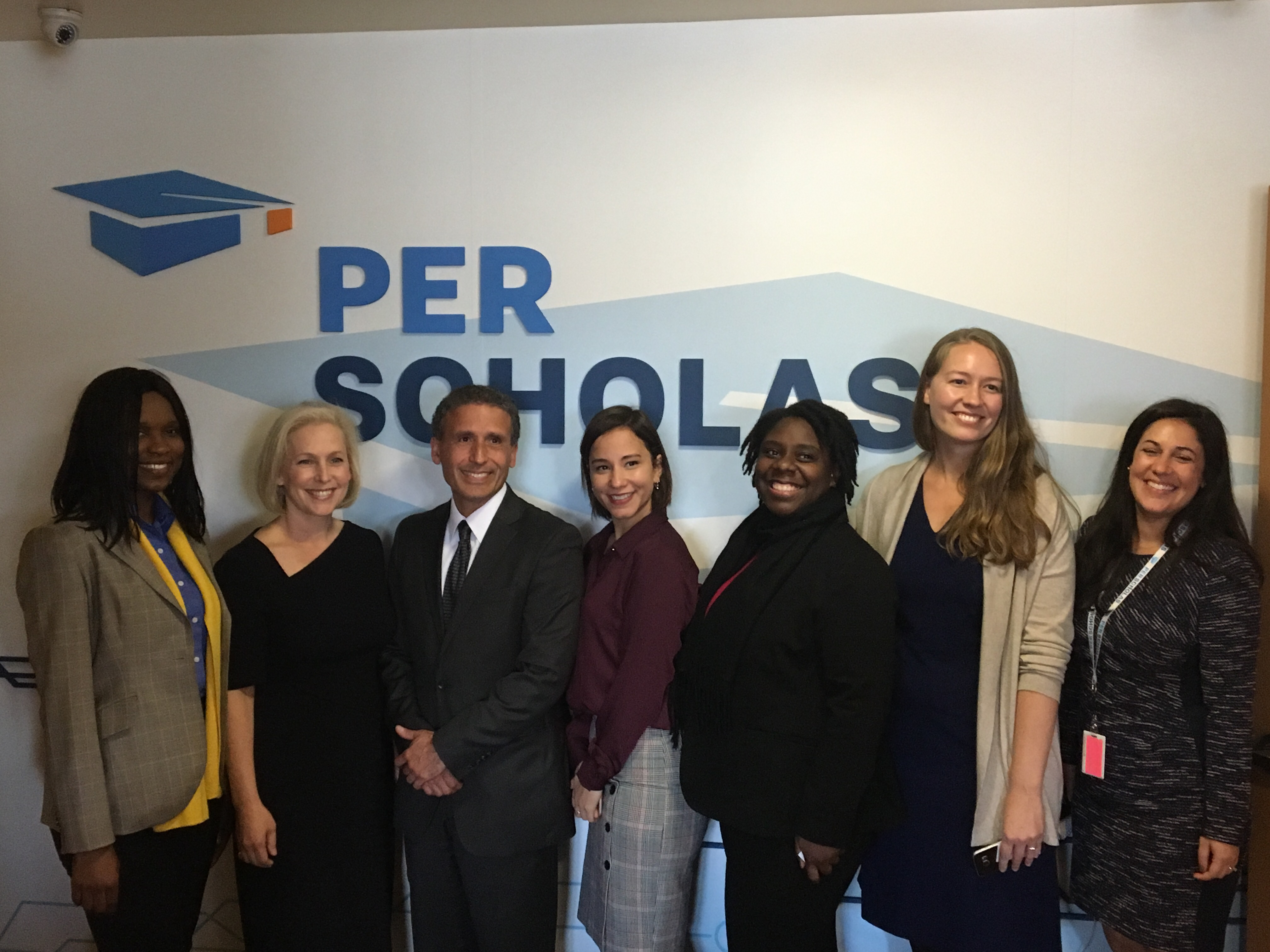 Seven smiling adults stand in a row in front of a wall with the Per Scholas logo, posing for a group photo.