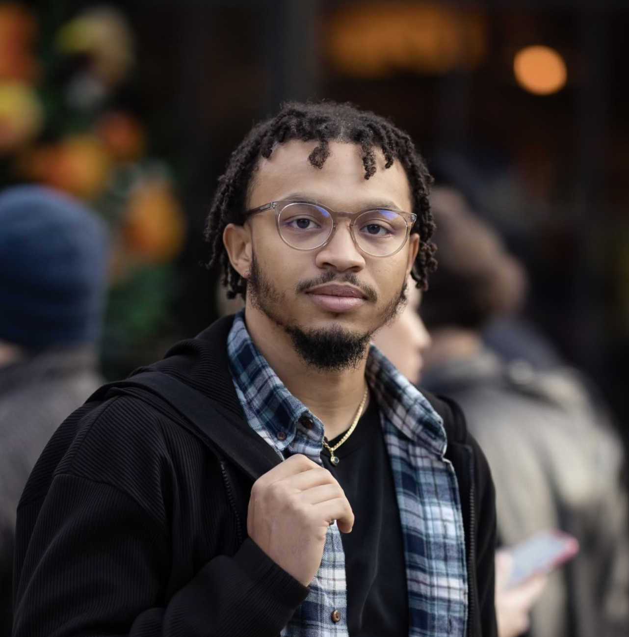 Young man standing outdoors looking at camera, holding a backpack strap.