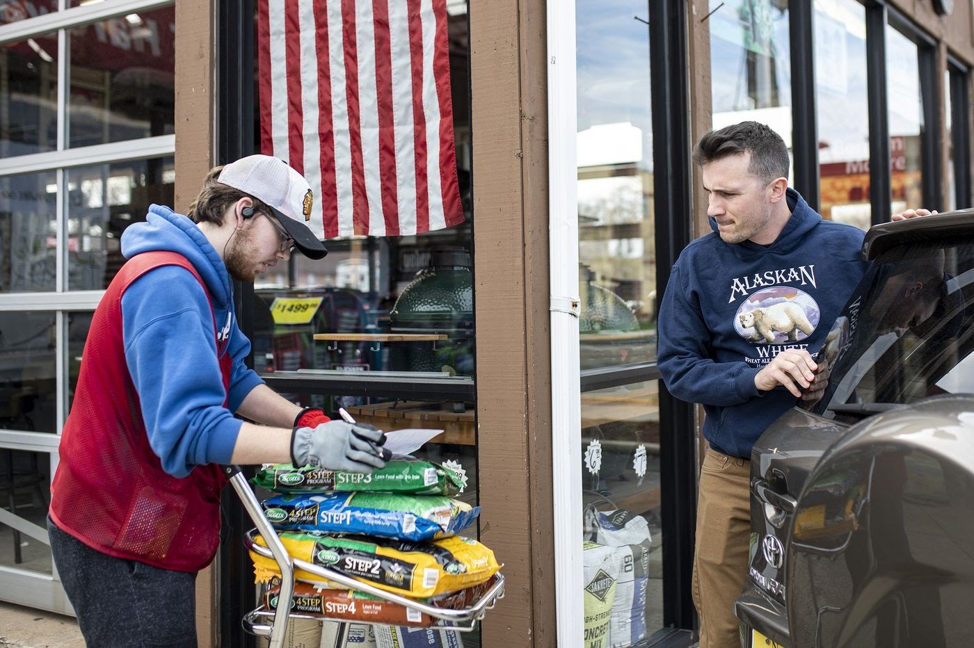 Worker loading bags of pet food onto a cart while a customer waits at a storefront doorway.