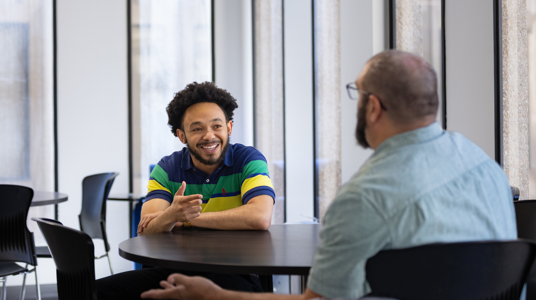 Young man in a striped polo shirt smiling and gesturing while talking with an older man in a denim shirt at a round table in a bright office.