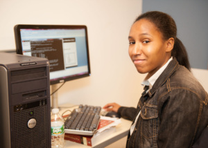 Young adult learner at a computer workstation, looking toward the camera while working on code shown on the monitor.