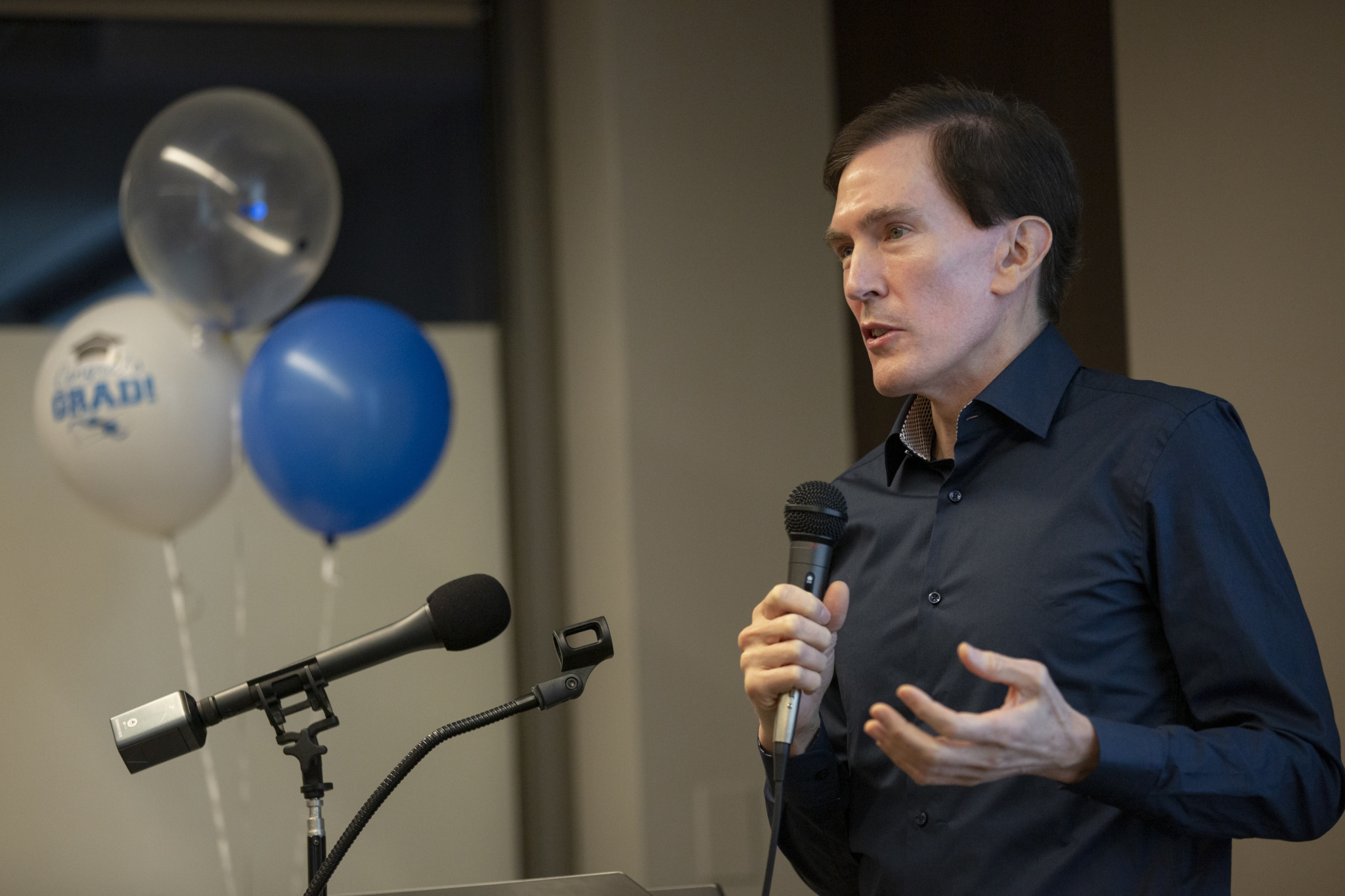 Man speaking into a microphone at a podium, gesturing with his free hand while balloons that read "Grad!" float in the background.