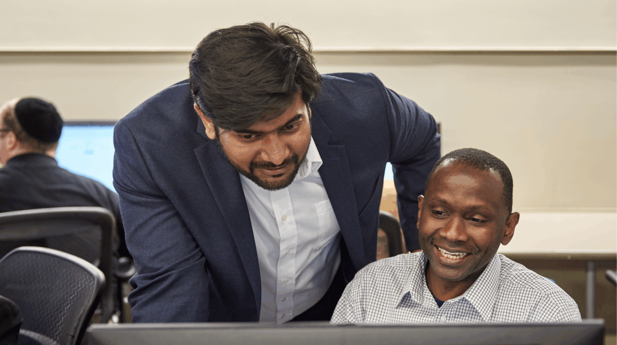 Instructor leaning over a learner's desk, both smiling as they look at a computer screen.