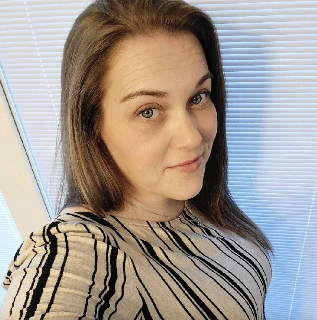 Headshot of Hope Shurba smiling while looking at the camera in front of a window with blinds.