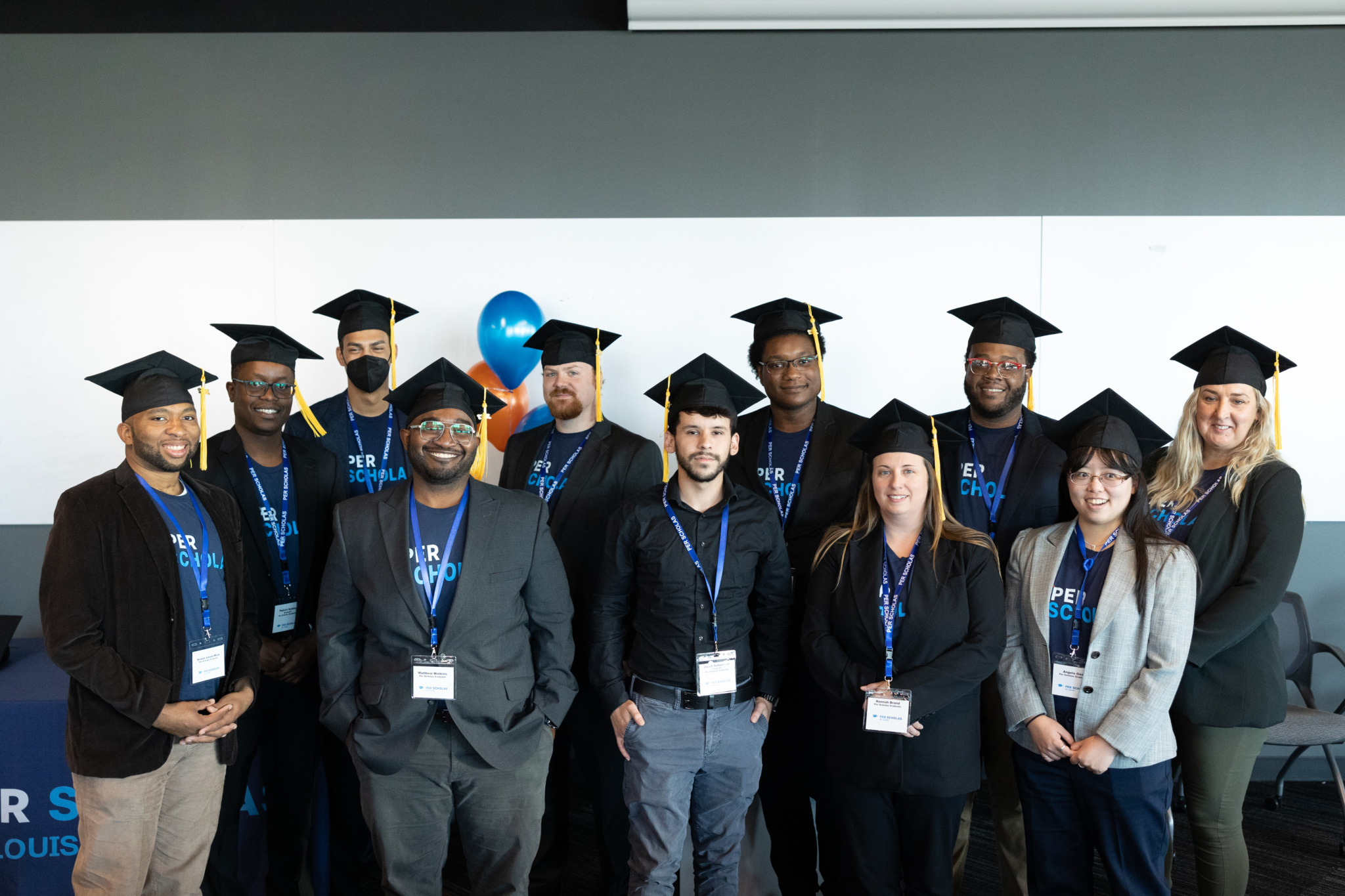 A diverse group of Per Scholas graduates wearing caps and name badges, smiling together at a graduation event.