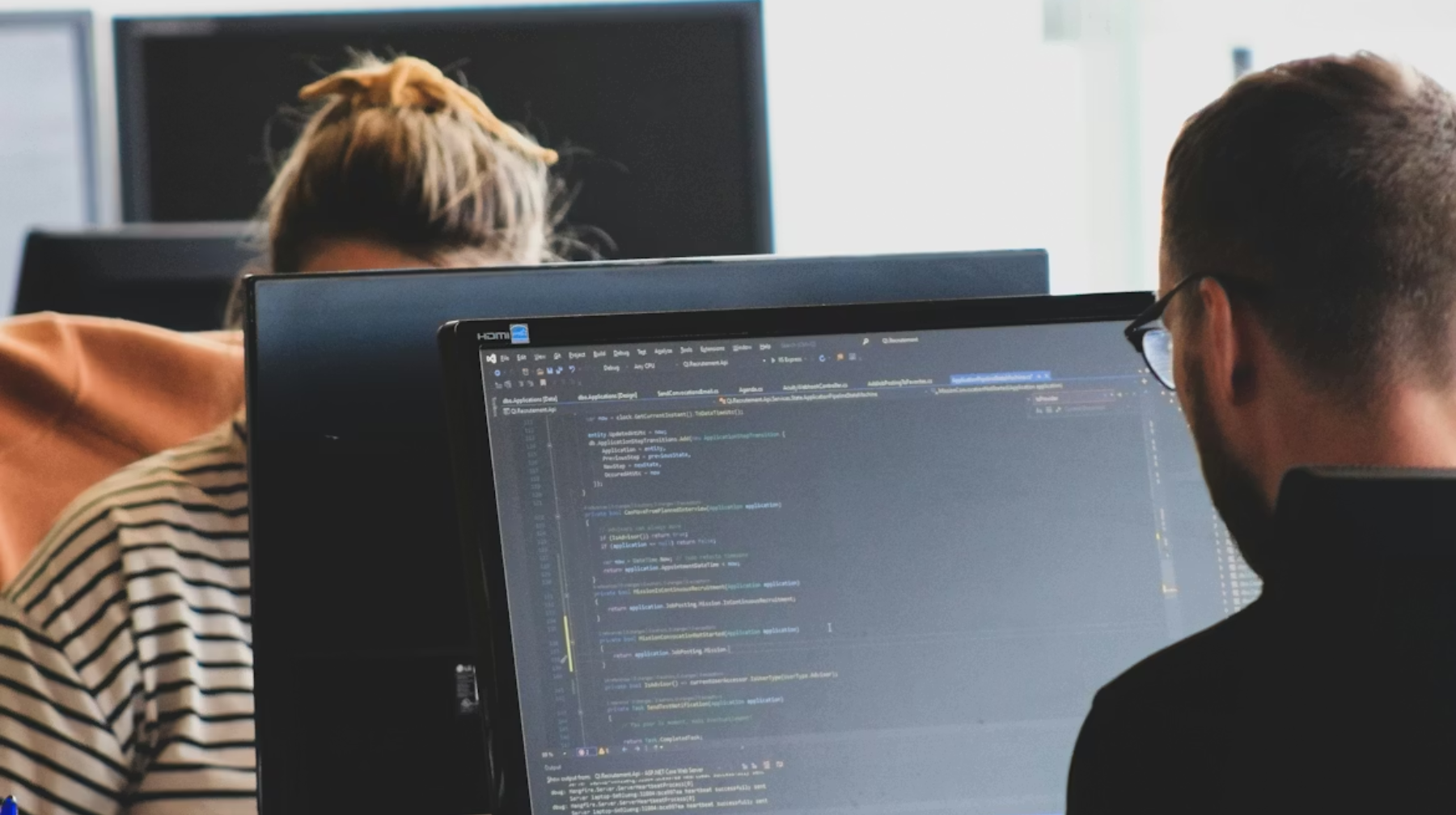 Two people working at computer monitors showing code, one person facing the screen and another partially visible behind a monitor.
