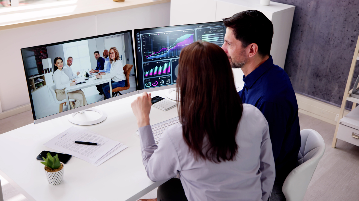 Male and female colleagues at a desk reviewing data on a monitor while joining a virtual meeting on a second screen.