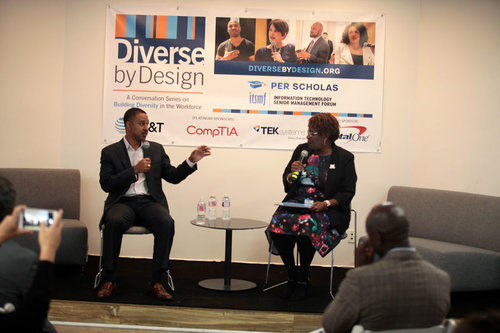 Two people seated on stage holding microphones in conversation in front of a "Diverse by Design" banner with sponsor logos.