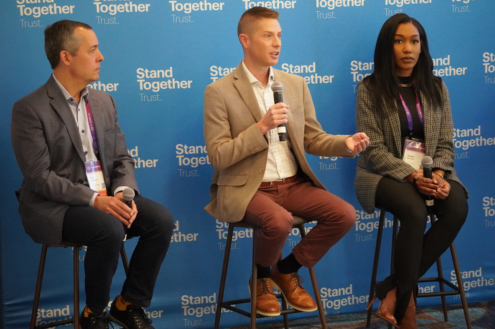 Three panelists seated on stools speaking at an event in front of a backdrop reading "Stand Together Trust."