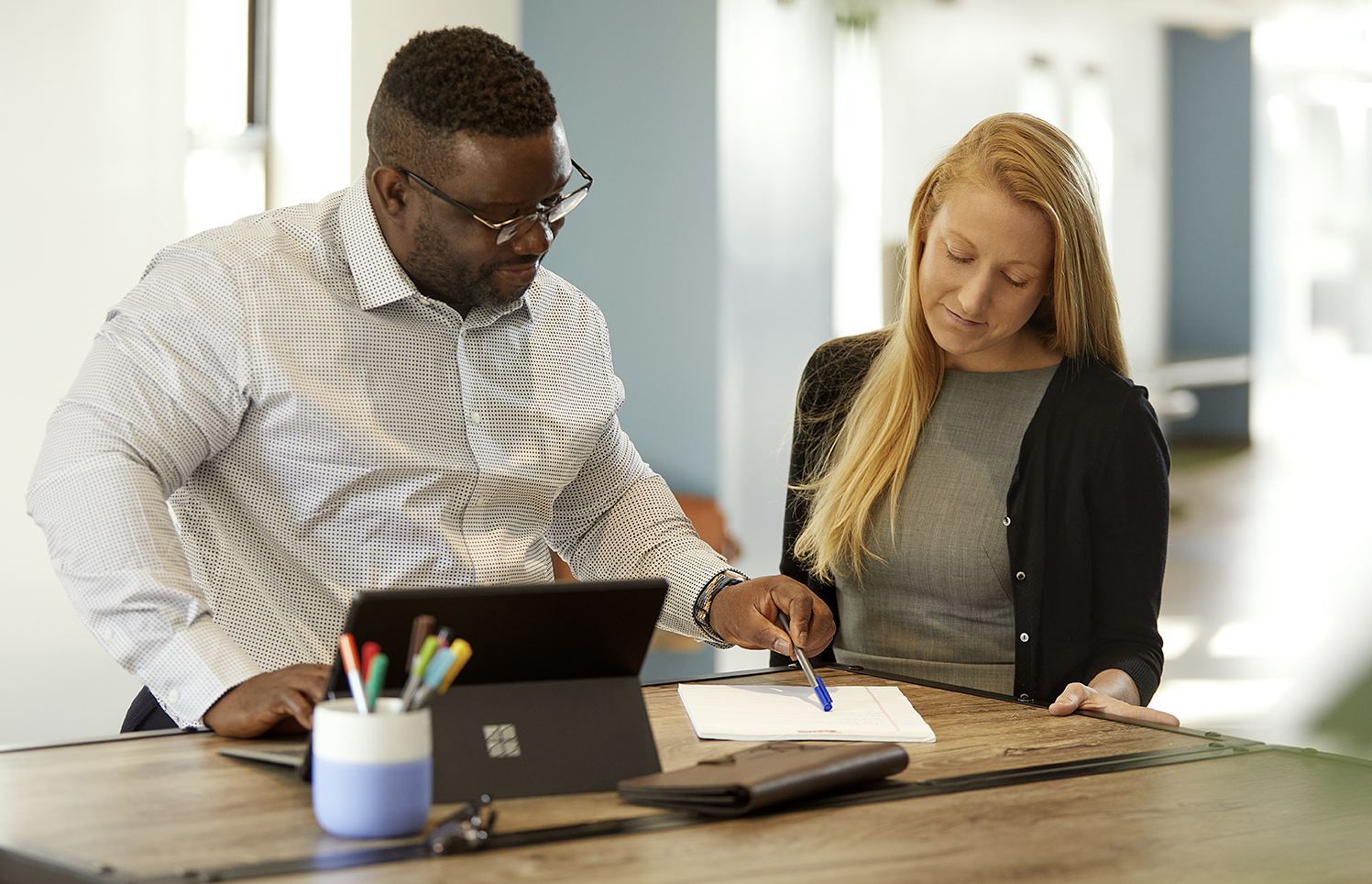 Two colleagues stand at a table in an office reviewing a document together, one pointing at the paper while the other listens.