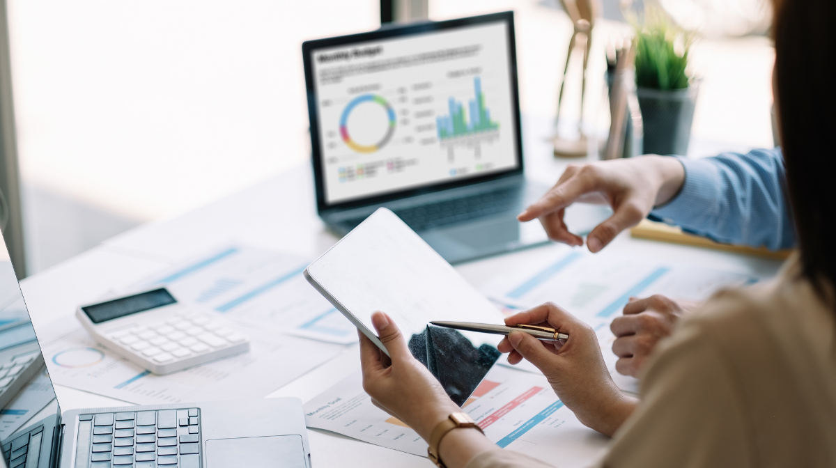 Two people review analytics at a desk; one points at a tablet while the other holds a stylus, with a laptop displaying graphs in the background.