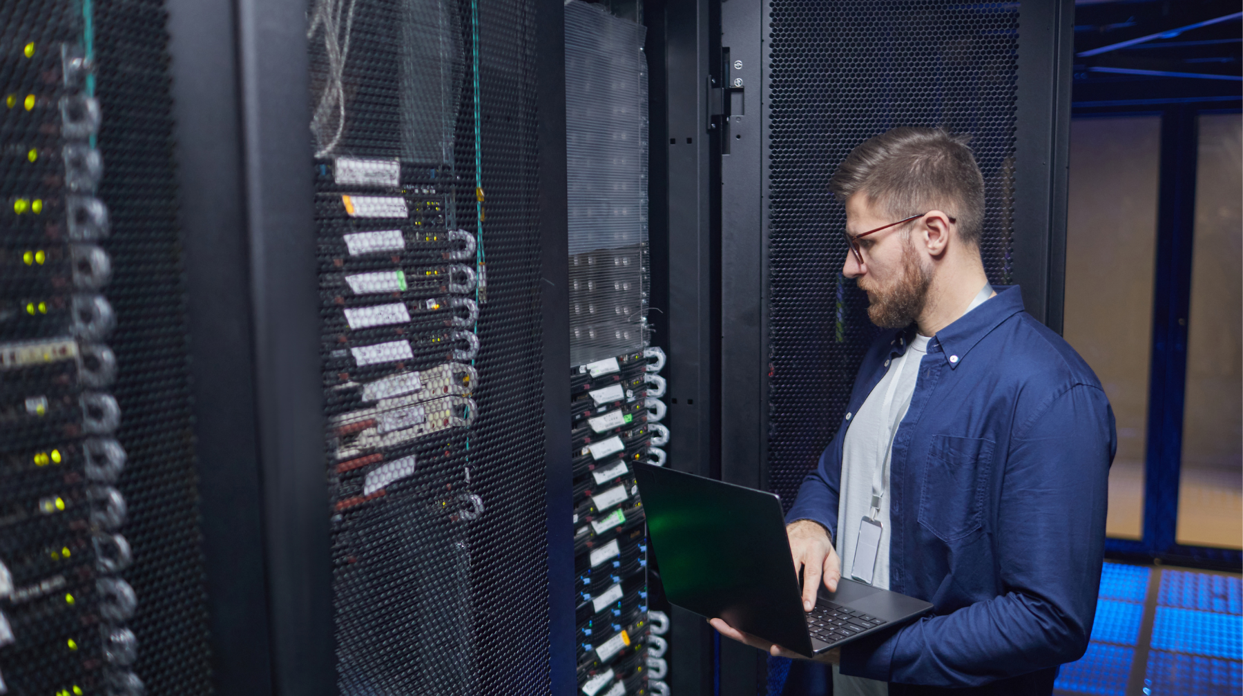 Technician standing in a server room checking a laptop while inspecting server racks.
