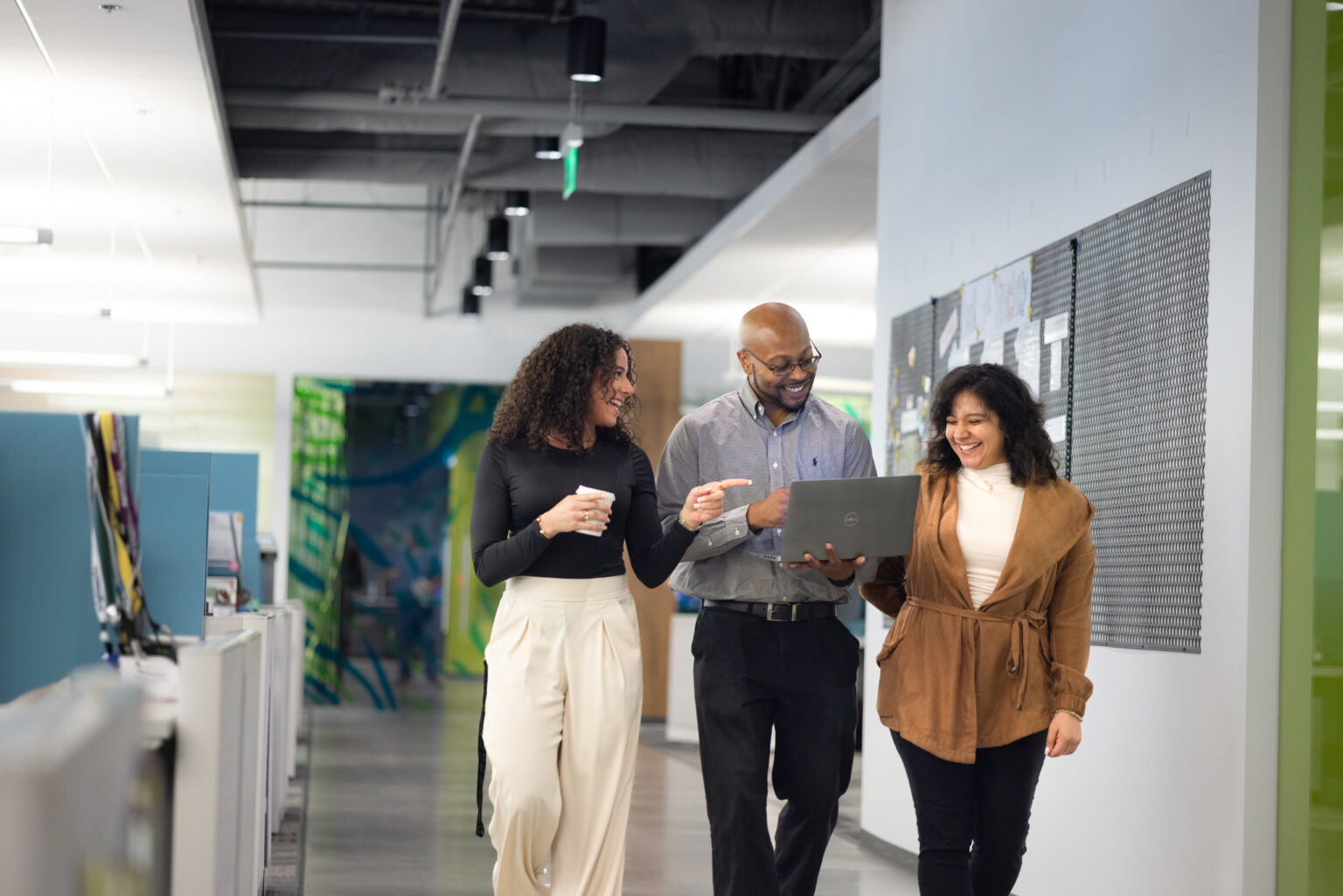 Three coworkers walking down an office hallway, smiling and discussing something on a laptop one person is holding.