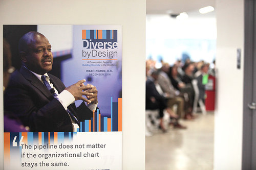 Poster reading "Diverse by Design" and quote "The pipeline does not matter if the organizational chart stays the same." with a blurred audience in a meeting room visible through a doorway.