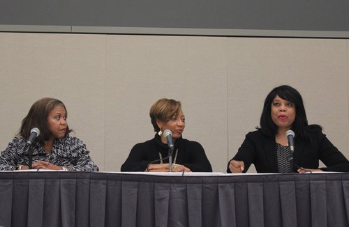Three women sit at a conference table with microphones, participating in a panel discussion.