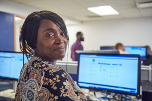 Adult learner at a computer in a classroom, looking toward the camera while classmates work in the background.