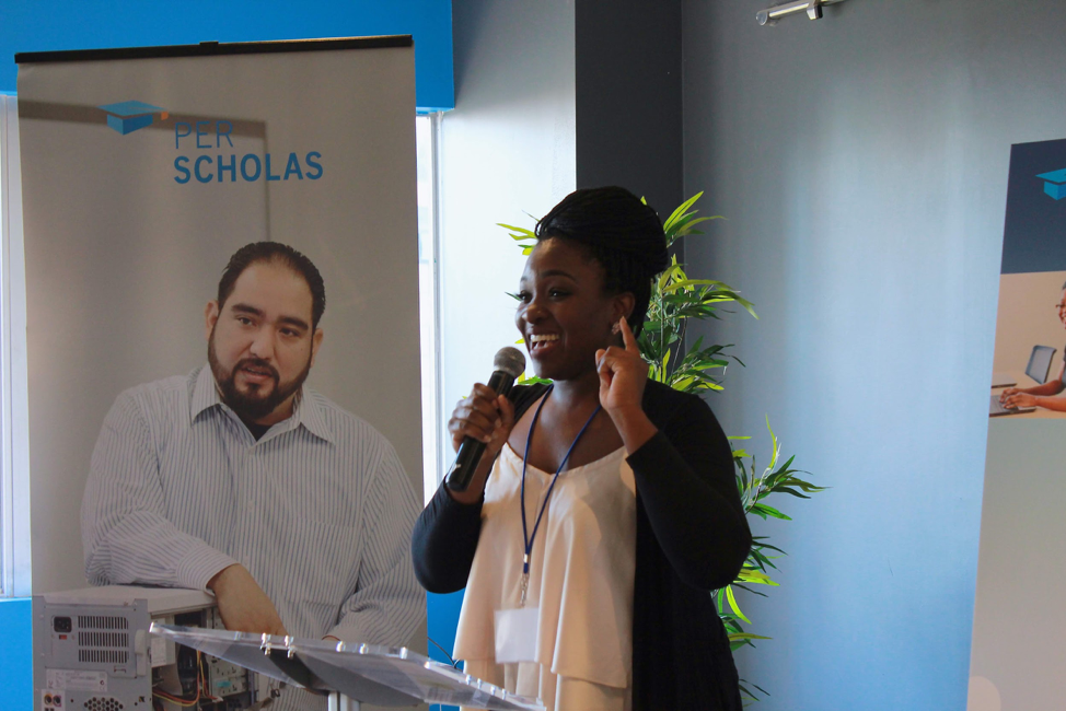 A woman speaking into a microphone at a Per Scholas event, smiling and gesturing while standing at a podium with Per Scholas banners behind her.