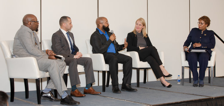 Five panelists sit on stage in a discussion; a man in the center holds a microphone while others listen.