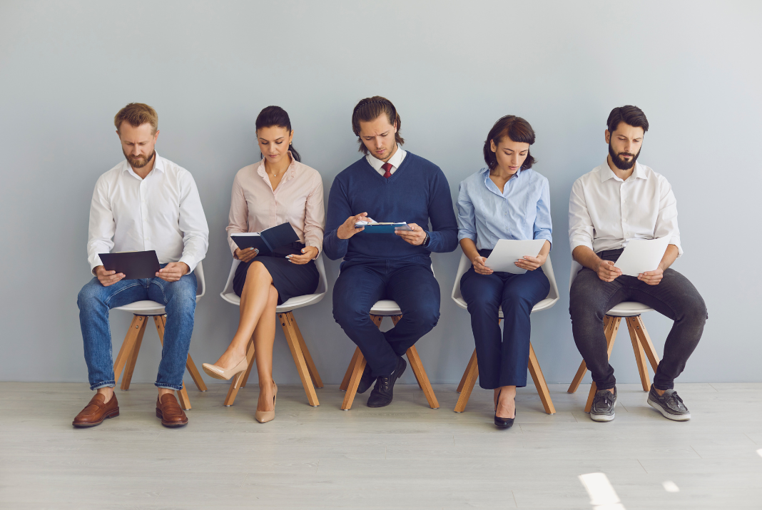 Five young professionals in business casual attire sitting side by side, each looking down at documents, or notebooks while waiting.