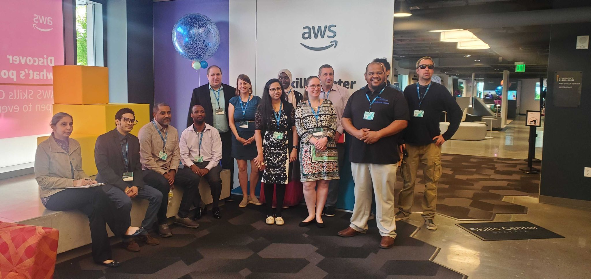 Group of a dozen adults standing and seated in an office lobby under an AWS sign, posing for a group photo.