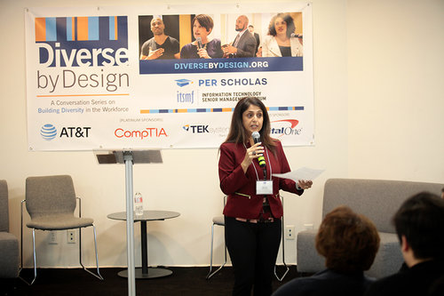 Woman speaker holding a microphone and notes addressing an audience in front of a "Diverse by Design" banner with sponsor logos.