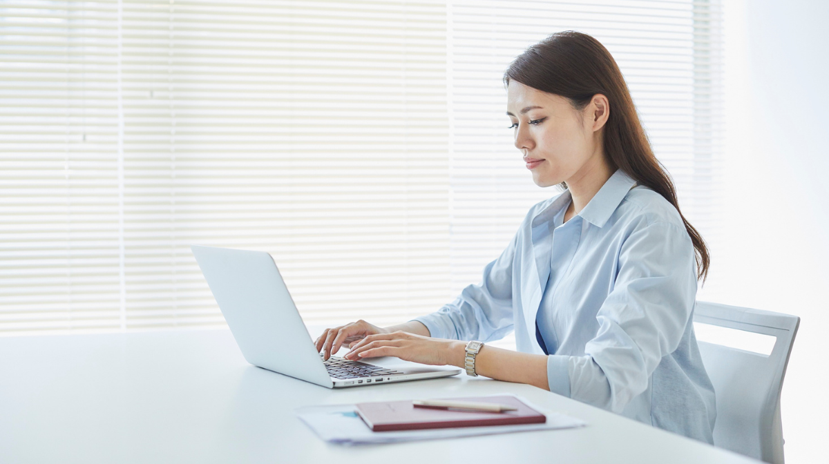 Woman working on a laptop at a bright desk with notebooks nearby.