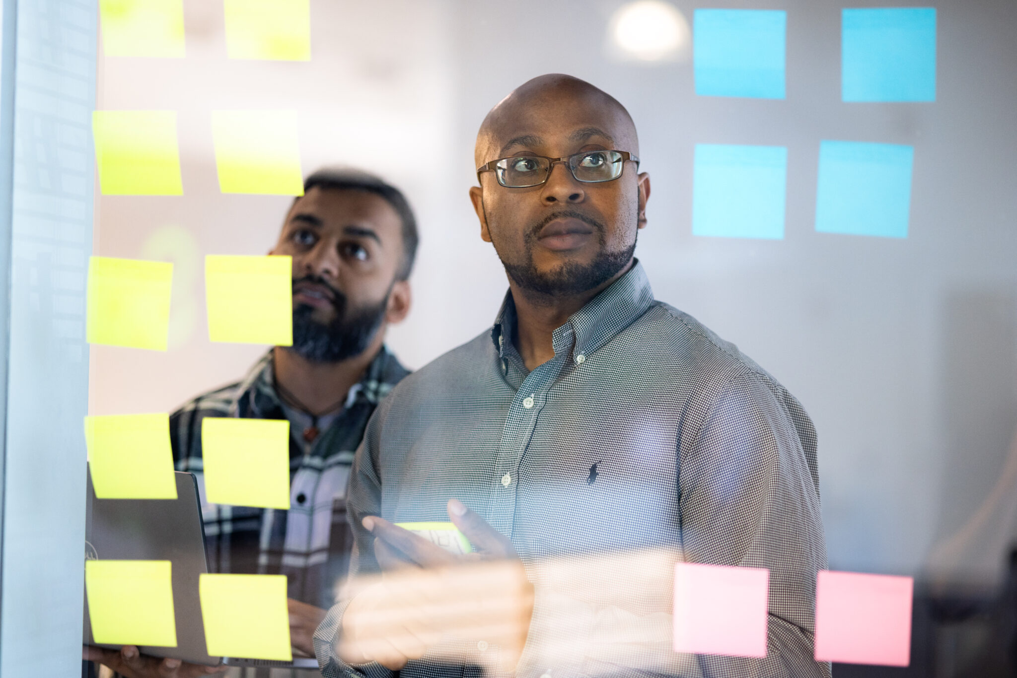 Two men standing behind a glass wall covered in sticky notes, looking thoughtfully at the notes.