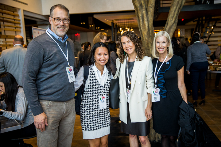 Four smiling adults wearing event badges pose together at a reception.