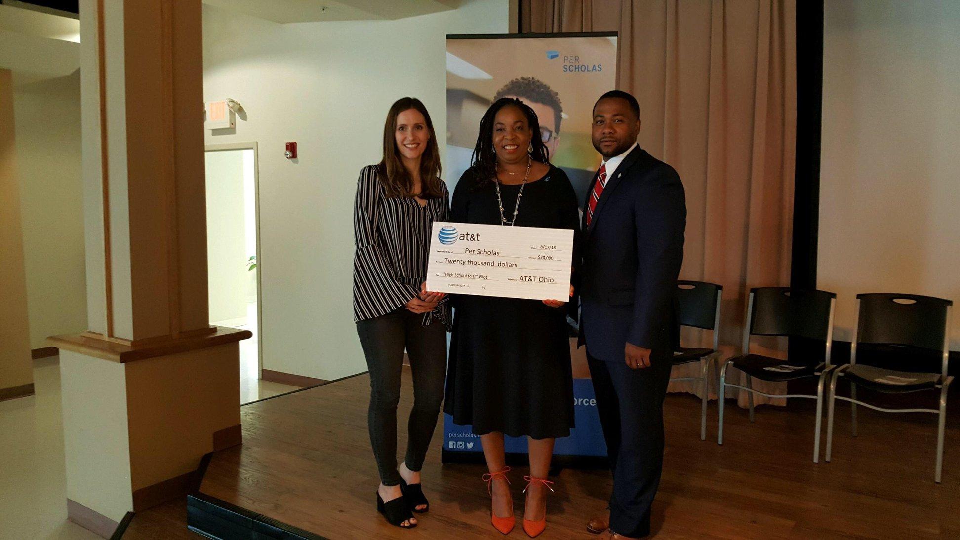 Three adults stand on a small stage holding a large ceremonial check for Per Scholas during a donation event.