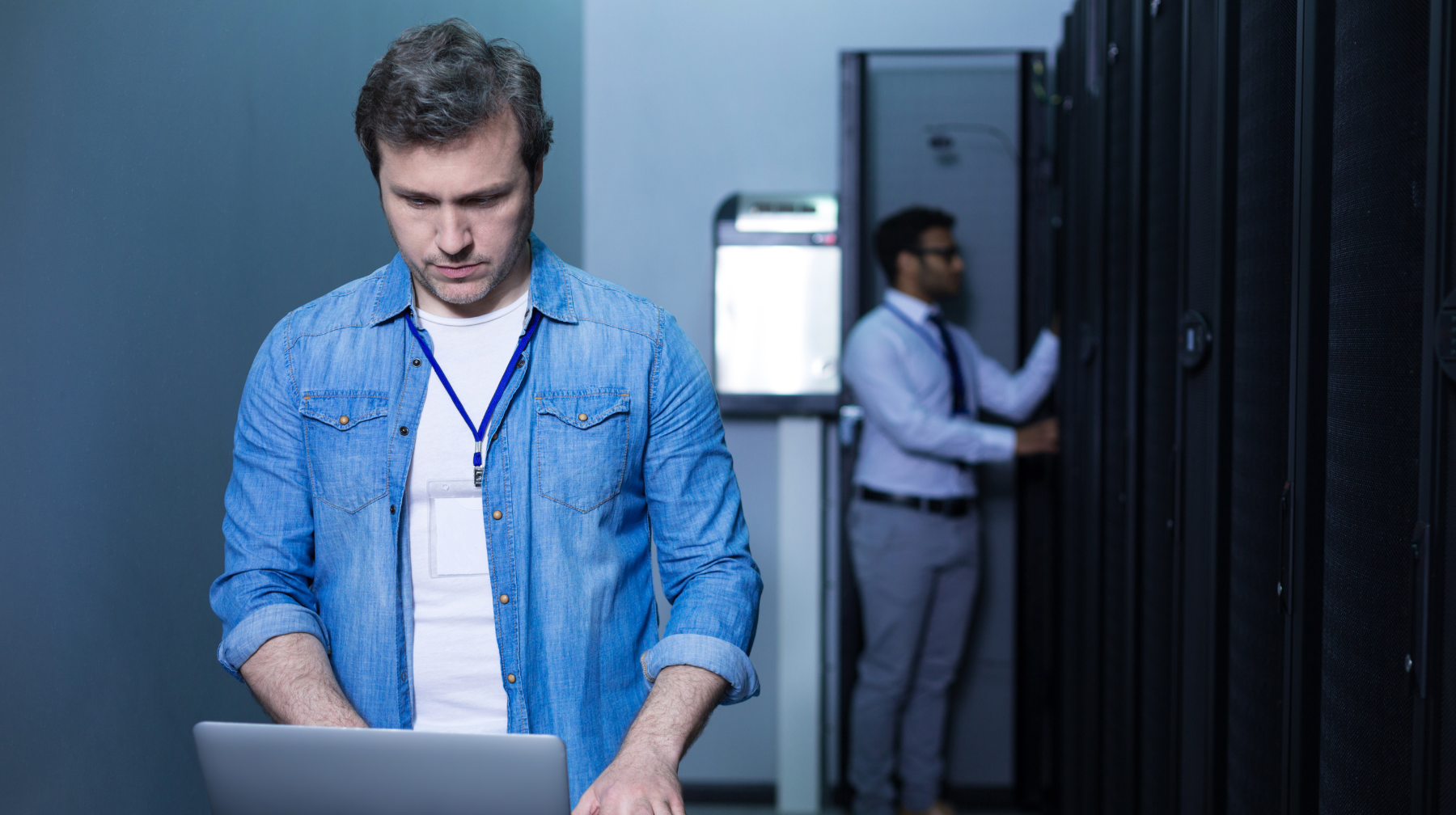 Two technicians working in a server room, one using a laptop in the foreground while another checks server racks in the background.