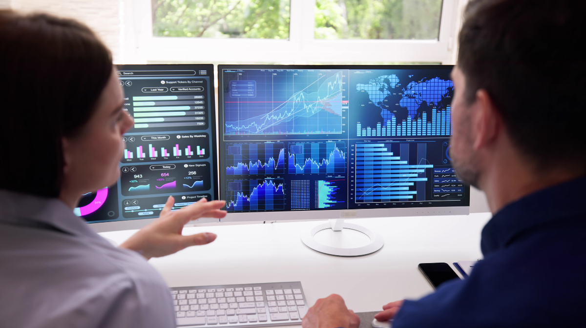Two colleagues at a desk discussing data visualizations on dual monitors showing charts, graphs and a world map.