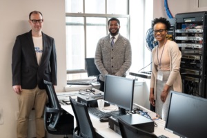 Three adult instructors stand smiling in a computer lab with workstations and networking equipment, posing for a group photo.