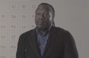Man speaking to camera in a classroom with a blurred periodic-table chart behind him.