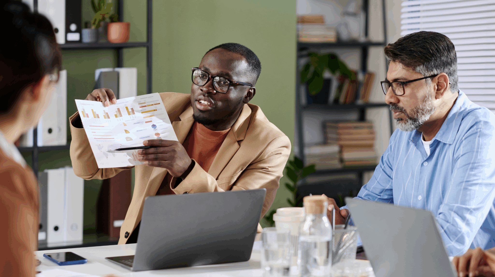Three colleagues review data at a conference table, one man holding up a printed chart to the others.
