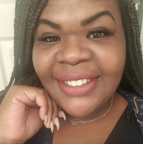 Kashena Miller smiling, with her hand near her face, wearing braided hair and a necklace.