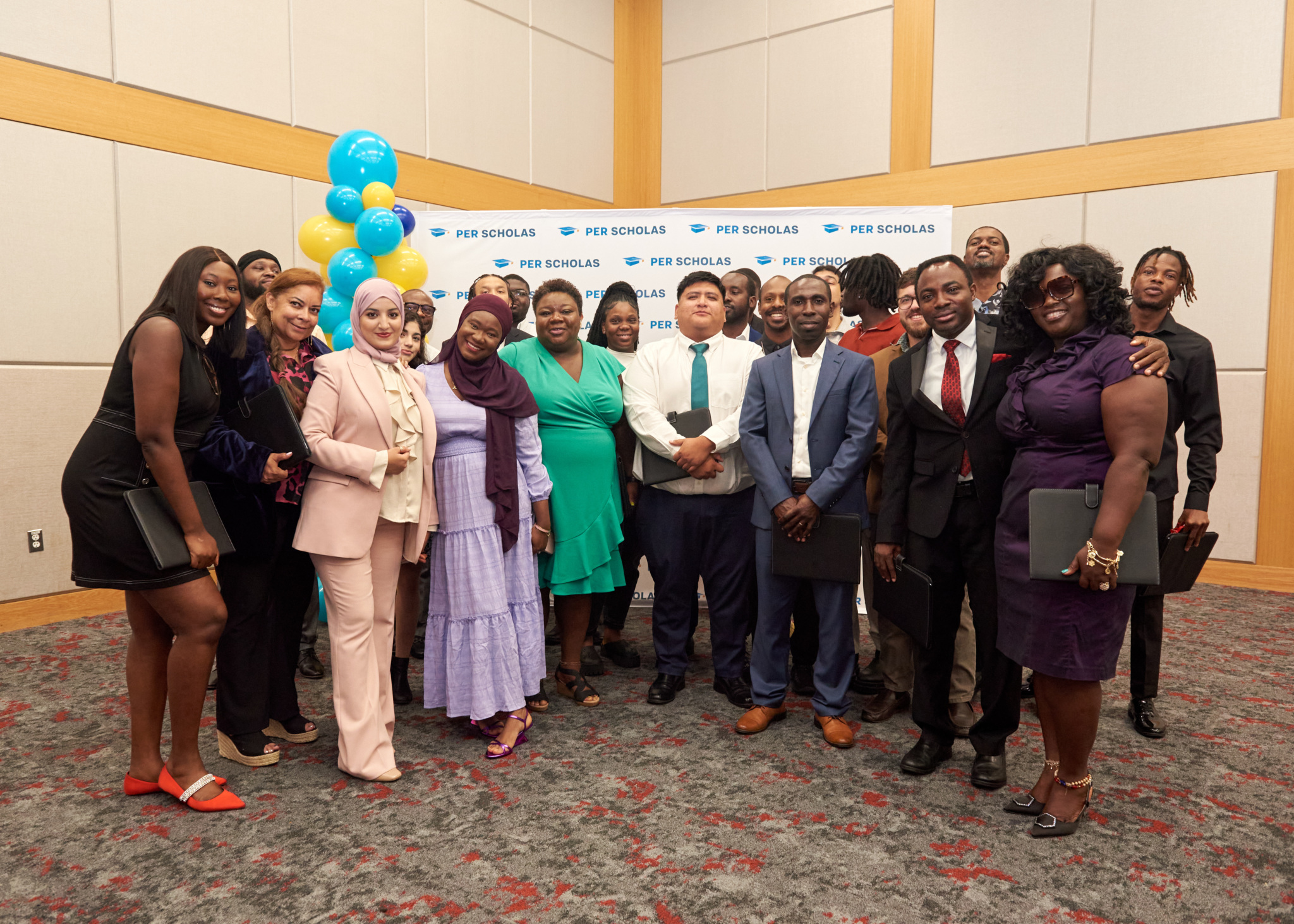 Group of smiling Per Scholas supporters posing together in a conference room with a Per Scholas step-and-repeat banner and balloon column behind them.