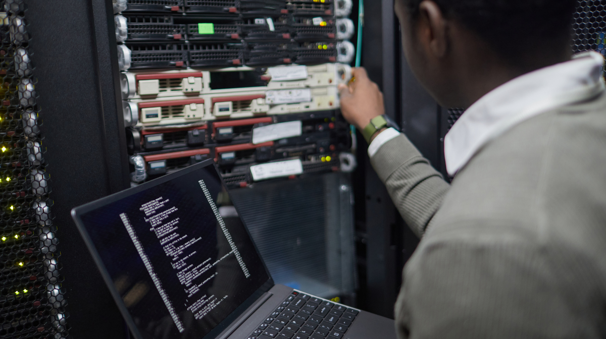 Person working at a server rack while monitoring code on a laptop.