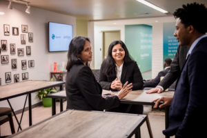 Three adult learners and an instructor talking and smiling around tall classroom tables in a Per Scholas learning space.