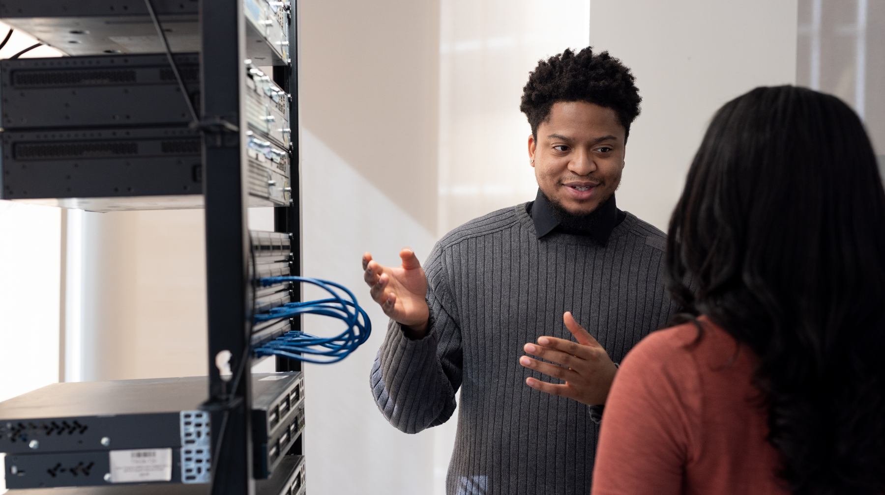 Two professionals talking beside a server rack, with one person explaining and gesturing to the other.