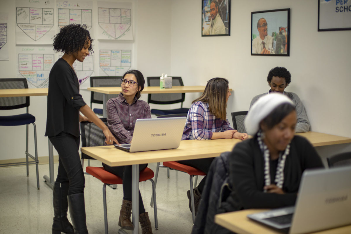 Instructor standing and pointing at a student’s laptop while a small group of adult learners work together in a classroom.