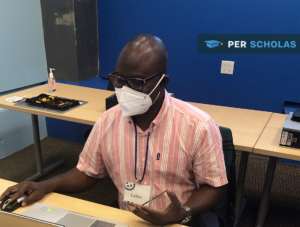 Man wearing a mask and name badge seated at a desk working on a laptop in a classroom.