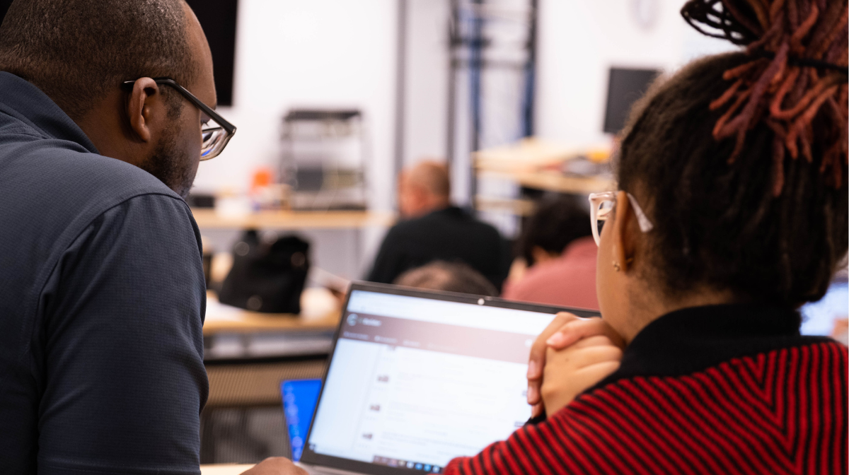 Two adult learners in a classroom collaborate over a laptop while other learners sit at desks in the background.