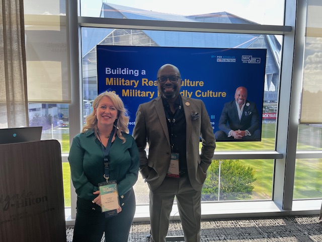 Two smiling adults wearing event badges stand in front of a screen that reads "Building a Military Ready Culture."