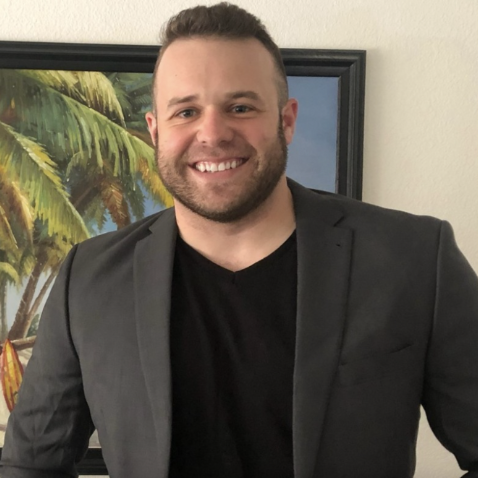 Headshot of Todd Theobald smiling in a blazer, standing in front of a framed painting of palm trees.